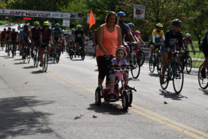 Mother assisting her daughter on an adapted trike with lots of participants in background.