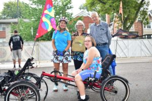 This is a photo of a woman on a recumbent tandem trike with her family/friends behind her. Everyone is happy and smiling.